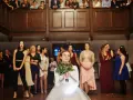 Panna młoda w białej sukni z bukietem zieleni, otoczona przez gości weselnych.