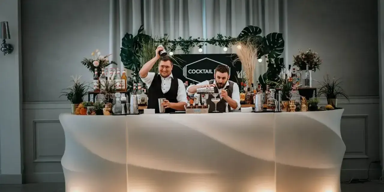 Dwóch barmanów przygotowuje drinki przy nowoczesnym, podświetlanym barze. To idealny drink bar na wesele, oferujący orzeźwiające napoje.