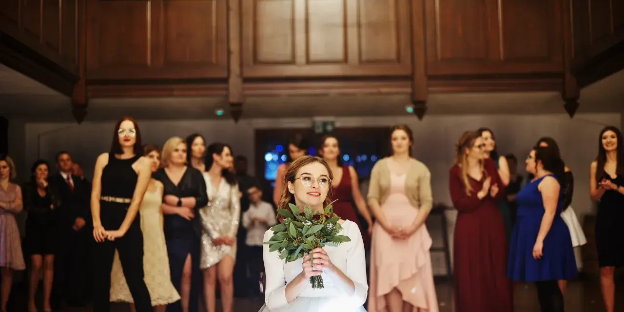 Panna młoda w białej sukni z bukietem zieleni, otoczona przez gości weselnych.