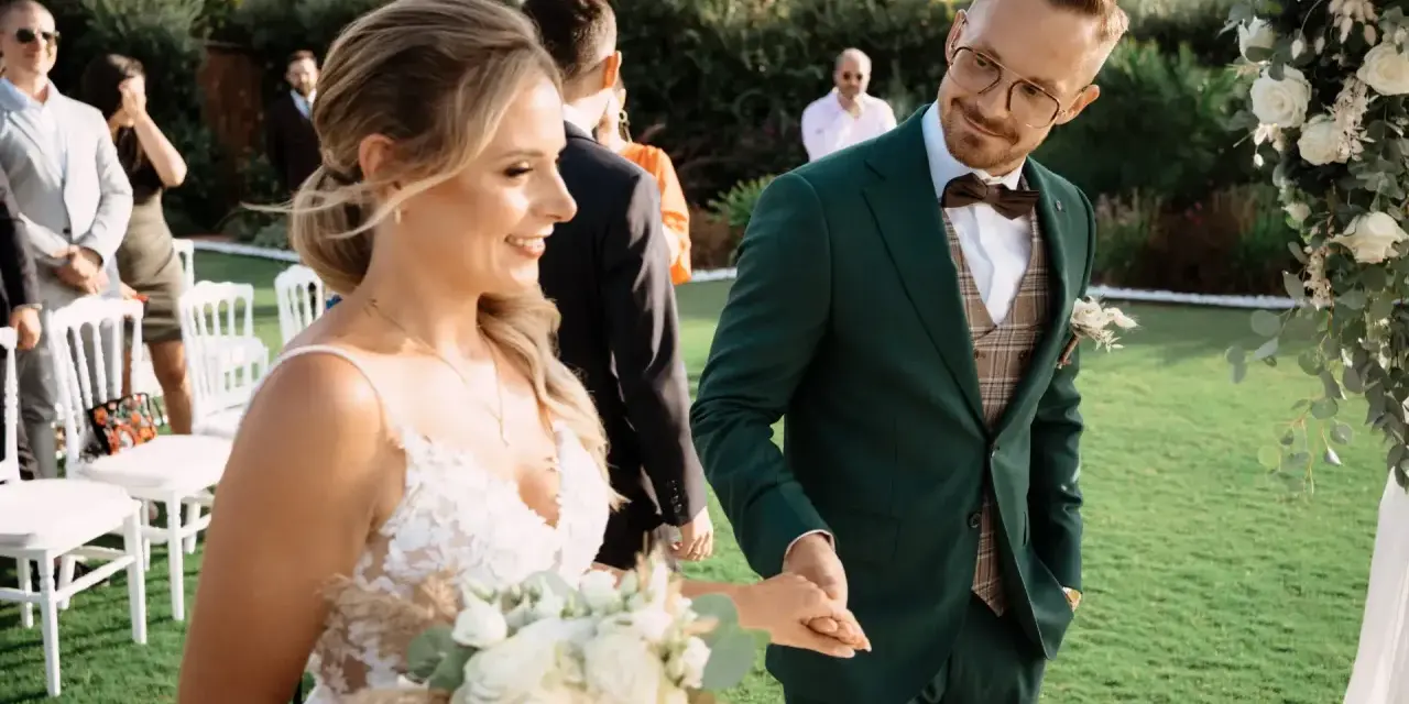 Para Młoda trzyma bukiet, a Pan Młody w zielonym garniturze uśmiecha się do niej. To piękny moment podczas urlopu okolicznościowego ślub.