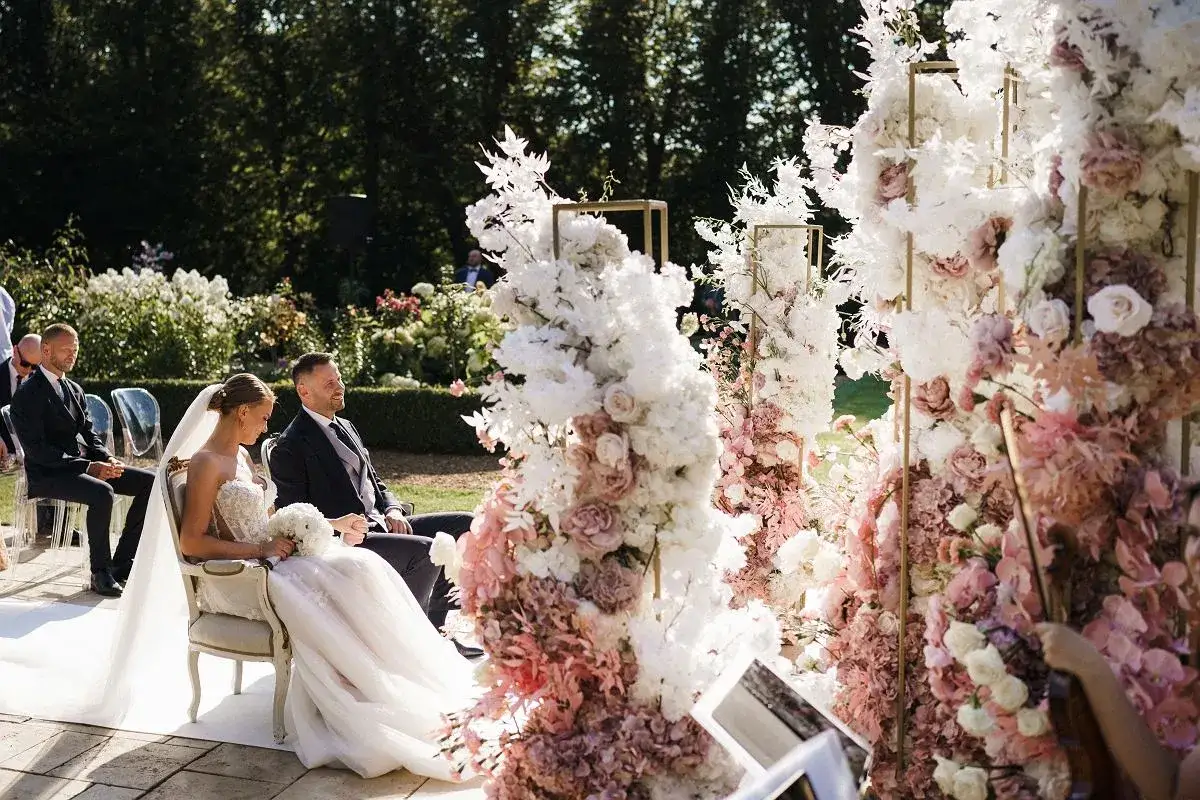 Para młoda siedzi na krzesłach podczas ceremonii. W tle bujne dekoracje kwiatowe. Taka oprawa to marzenie, warto sprawdzić wedding planner cena.