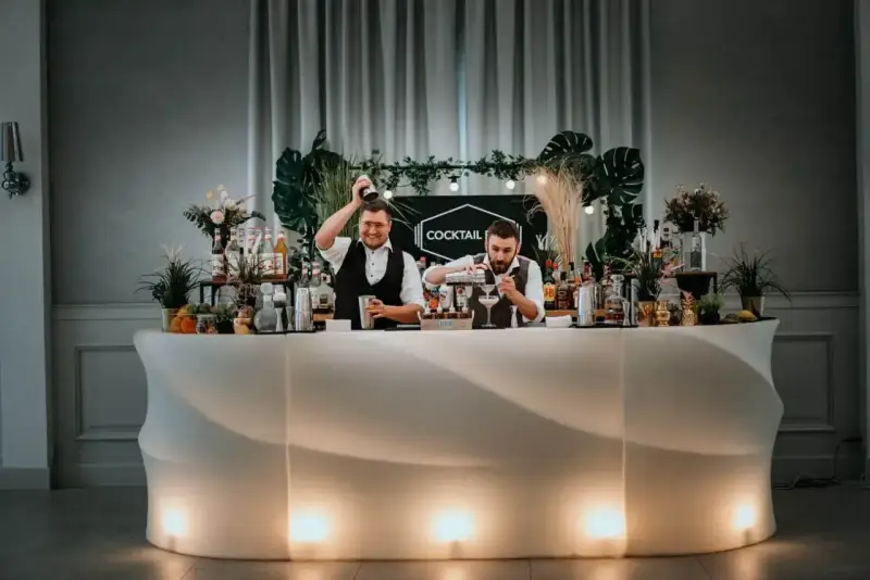 Dwóch barmanów przygotowuje drinki przy nowoczesnym, podświetlanym barze. To idealny drink bar na wesele, oferujący orzeźwiające napoje.